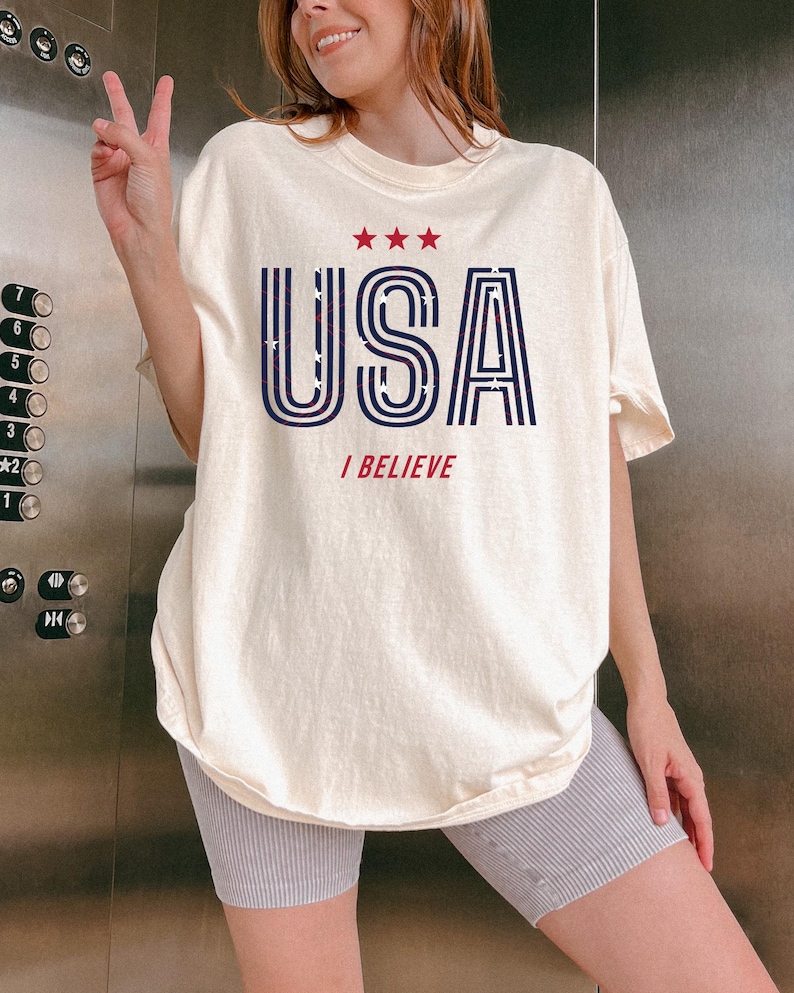 May include: Cream-colored t-shirt with the word "USA" in navy blue, outlined in white, and filled with a star pattern. Above the word are three red stars, and below it, the words "I BELIEVE" in red.