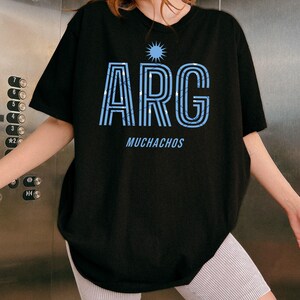 May include: Black t-shirt with the word "ARG" in light blue, featuring a sunburst design above and the word "MUCHACHOS" below. The letters have a striped pattern. The shirt is a casual style.