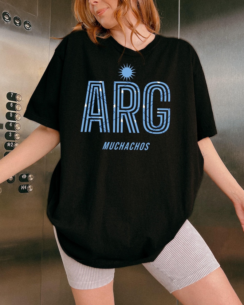 May include: Black t-shirt with the word "ARG" in light blue, featuring a sunburst design above and the word "MUCHACHOS" below. The letters have a striped pattern. The shirt is a casual style.