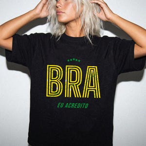 May include: Black t-shirt with the word "BRA" in yellow outlined letters with a green and yellow design. Above the word are five green stars. Below the word is the text "EU ACREDITO" in green.