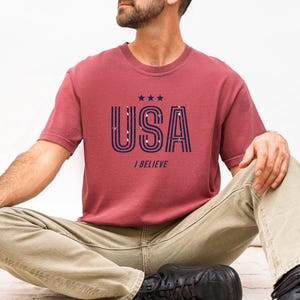 May include: A maroon t-shirt with the word "USA" in navy blue, featuring a striped design and three stars above the text. Below the text is the phrase "I BELIEVE". The shirt is paired with khaki pants.