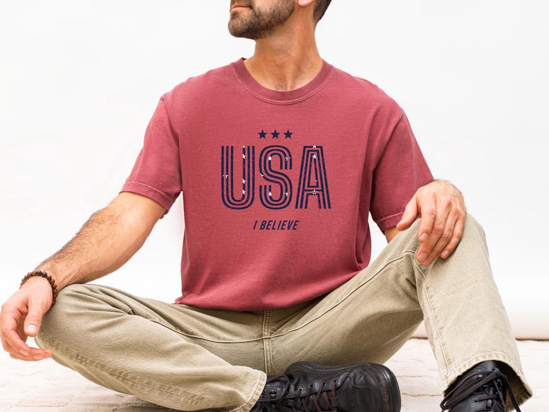 May include: A maroon t-shirt with the word "USA" in navy blue, featuring a striped design and three stars above the text. Below the text is the phrase "I BELIEVE". The shirt is paired with khaki pants.