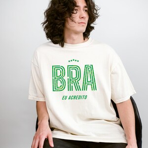 May include: Cream-colored t-shirt with the word "BRA" in green, outlined with a striped design. Below the word "BRA" is the phrase "EU ACREDITO" in green. The shirt is worn by a person.