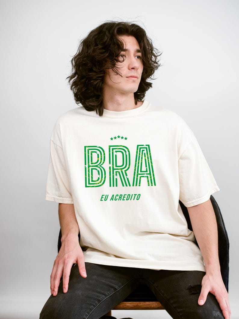 May include: Cream-colored t-shirt with the word "BRA" in green, outlined with a striped design. Below the word "BRA" is the phrase "EU ACREDITO" in green. The shirt is worn by a person.