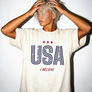 May include: Cream-colored t-shirt with the word "USA" in navy blue and red, with three red stars above the letters. Below the word "USA" is the phrase "I BELIEVE" in red. The shirt has a classic crew neck and short sleeves.