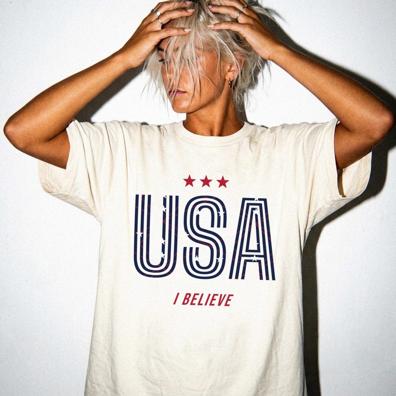 May include: Cream-colored t-shirt with the word "USA" in navy blue and red, with three red stars above the letters. Below the word "USA" is the phrase "I BELIEVE" in red. The shirt has a classic crew neck and short sleeves.