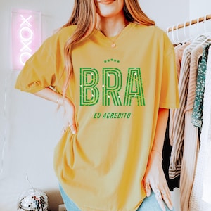 May include: Mustard yellow t-shirt with the word "BRA" in green, outlined letters, and the text "EU ACREDITO" below. The shirt has a crew neck and short sleeves. The shirt is a relaxed fit.