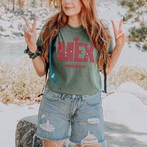 May include: Green t-shirt with the word "M&Eacute;X" in red block letters and the words "VAMOS MEXICO" below. The shirt is paired with light blue denim shorts. The image is taken outdoors.