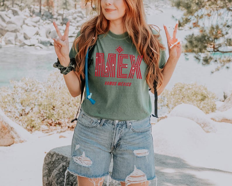 May include: Green t-shirt with the word "M&Eacute;X" in red block letters and the words "VAMOS MEXICO" below. The shirt is paired with light blue denim shorts. The image is taken outdoors.