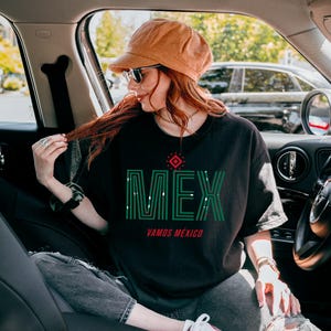May include: Black t-shirt with the word "MEX" in green, with a red and white design above it and the words "VAMOS M&Eacute;XICO" below. The shirt is worn with a brown hat and sunglasses.