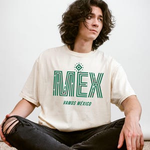 May include: Cream-colored t-shirt with the word "MEX" in green, featuring a geometric design and the phrase "VAMOS M&Eacute;XICO." The shirt is worn with black jeans. The overall theme is Mexican pride.