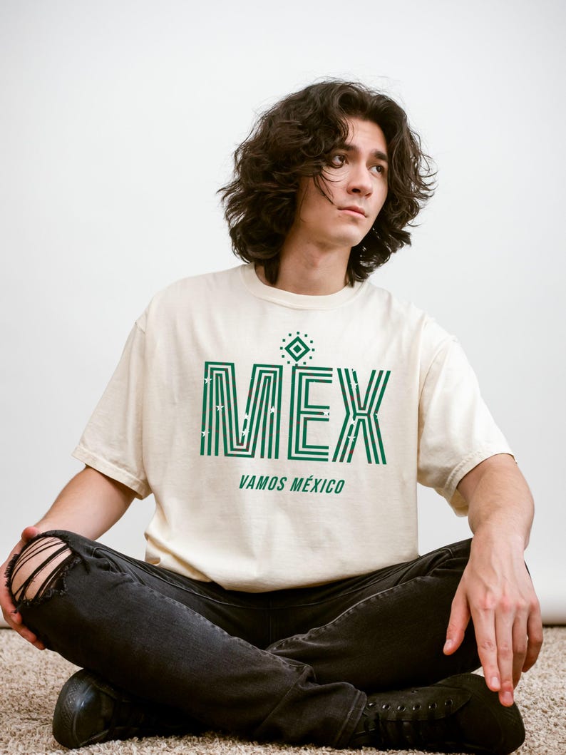 May include: Cream-colored t-shirt with the word "MEX" in green, featuring a geometric design and the phrase "VAMOS M&Eacute;XICO." The shirt is worn with black jeans. The overall theme is Mexican pride.