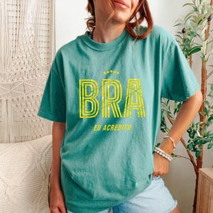 May include: Teal-colored t-shirt with the word "BRA" in yellow, featuring a striped design. Above the word are three yellow stars. Below the word is the text "EU ACREDITO".