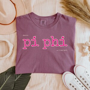 Könnte beinhalten: Ein malvenfarbenes Kurzarm-T-Shirt mit dem Text "she's a pi phi" in einer kräftigen rosa Schrift mit weißer Umrandung und dem kleineren Text "...i can tell" darunter in Weiß. Das T-Shirt hat einen Rundhalsausschnitt und eine Standardpassform.