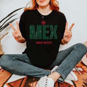 May include: Black t-shirt with the word "MEX" in green, with a red accent and the words "VAMOS M&Eacute;XICO" underneath. The person is wearing blue jeans and white sneakers. A straw hat and a potted plant are in the background.