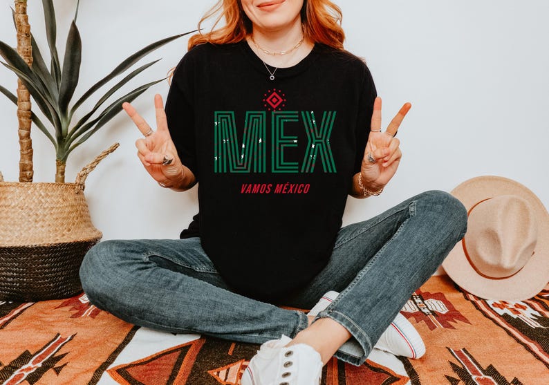 May include: Black t-shirt with the word "MEX" in green, with a red accent and the words "VAMOS M&Eacute;XICO" underneath. The person is wearing blue jeans and white sneakers. A straw hat and a potted plant are in the background.