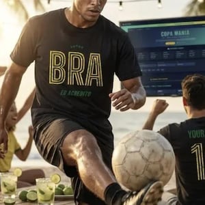 May include: A black t-shirt with the word "BRA" in yellow, with the text "EU ACREDITO" underneath. A soccer player is kicking a soccer ball on a beach. The background includes a screen with the words "COPA MANIA".
