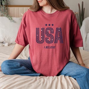 May include: A maroon-colored t-shirt with the word "USA" in navy blue, featuring a star design and the phrase "I BELIEVE". The shirt is a casual style, suitable for everyday wear.
