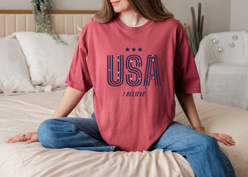 May include: A maroon-colored t-shirt with the word "USA" in navy blue, featuring a star design and the phrase "I BELIEVE". The shirt is a casual style, suitable for everyday wear.