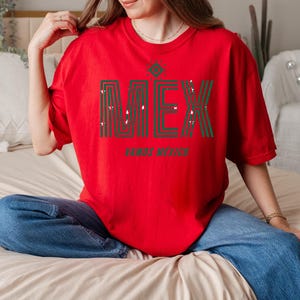 May include: A red t-shirt with the word "MEX" in green and white, with a decorative design above the letters. Below the word "MEX" is the phrase "VAMOS MEXICO". The shirt is worn with blue jeans.