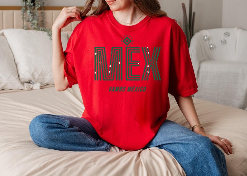 May include: A red t-shirt with the word "MEX" in green and white, with a decorative design above the letters. Below the word "MEX" is the phrase "VAMOS MEXICO". The shirt is worn with blue jeans.