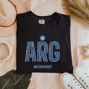 May include: Black t-shirt with the word "ARG" in light blue, with a sun graphic above and the word "MUCHACHOS" below. The shirt is surrounded by a hat, headband, shoes, and other accessories.