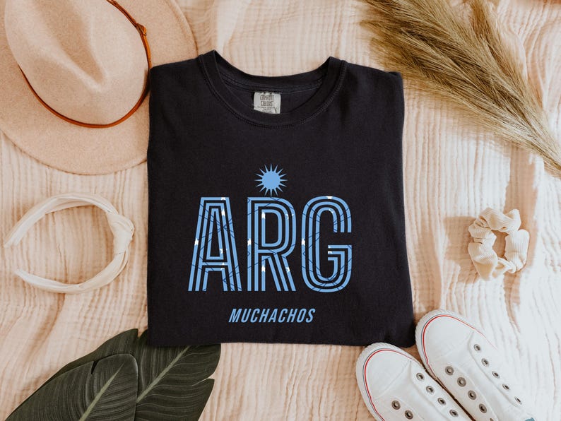 May include: Black t-shirt with the word "ARG" in light blue, with a sun graphic above and the word "MUCHACHOS" below. The shirt is surrounded by a hat, headband, shoes, and other accessories.