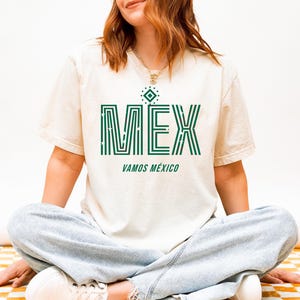May include: Cream-colored t-shirt with the word "MEX" in green, featuring a geometric design. Below, the text "VAMOS M&Eacute;XICO" is also in green. The shirt is worn by a person sitting cross-legged.
