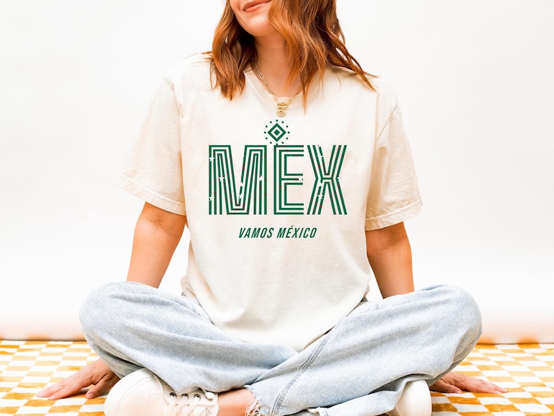 May include: Cream-colored t-shirt with the word "MEX" in green, featuring a geometric design. Below, the text "VAMOS M&Eacute;XICO" is also in green. The shirt is worn by a person sitting cross-legged.