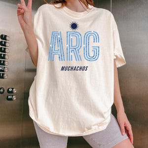 May include: Cream-colored t-shirt with the word "ARG" in light blue, outlined in a darker blue, and filled with white stars. Below the word "ARG" is the word "MUCHACHOS" in a dark blue font. A navy blue sunburst design is above the word "ARG".