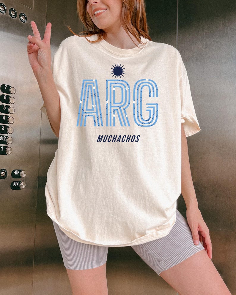 May include: Cream-colored t-shirt with the word "ARG" in light blue, outlined in a darker blue, and filled with white stars. Below the word "ARG" is the word "MUCHACHOS" in a dark blue font. A navy blue sunburst design is above the word "ARG".