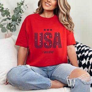 May include: Red t-shirt with the word "USA" in navy blue and white, with three stars above and the words "I BELIEVE" below. The shirt is worn with blue jeans.