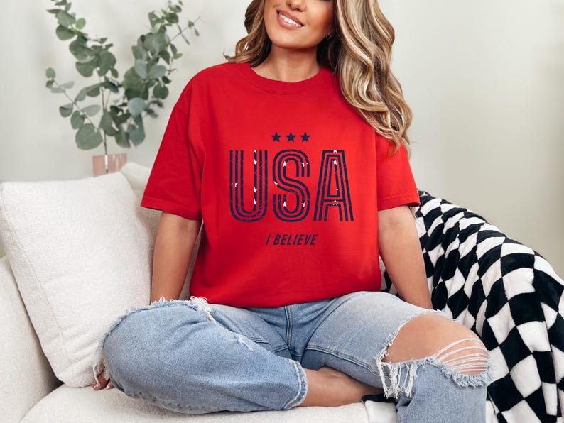 May include: Red t-shirt with the word "USA" in navy blue and white, with three stars above and the words "I BELIEVE" below. The shirt is worn with blue jeans.