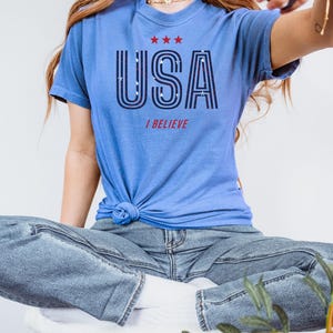 May include: A light blue t-shirt with the word "USA" in a bold, outlined font, and the words "I BELIEVE" below it. The shirt has three red stars above the word "USA". The shirt is knotted at the waist.