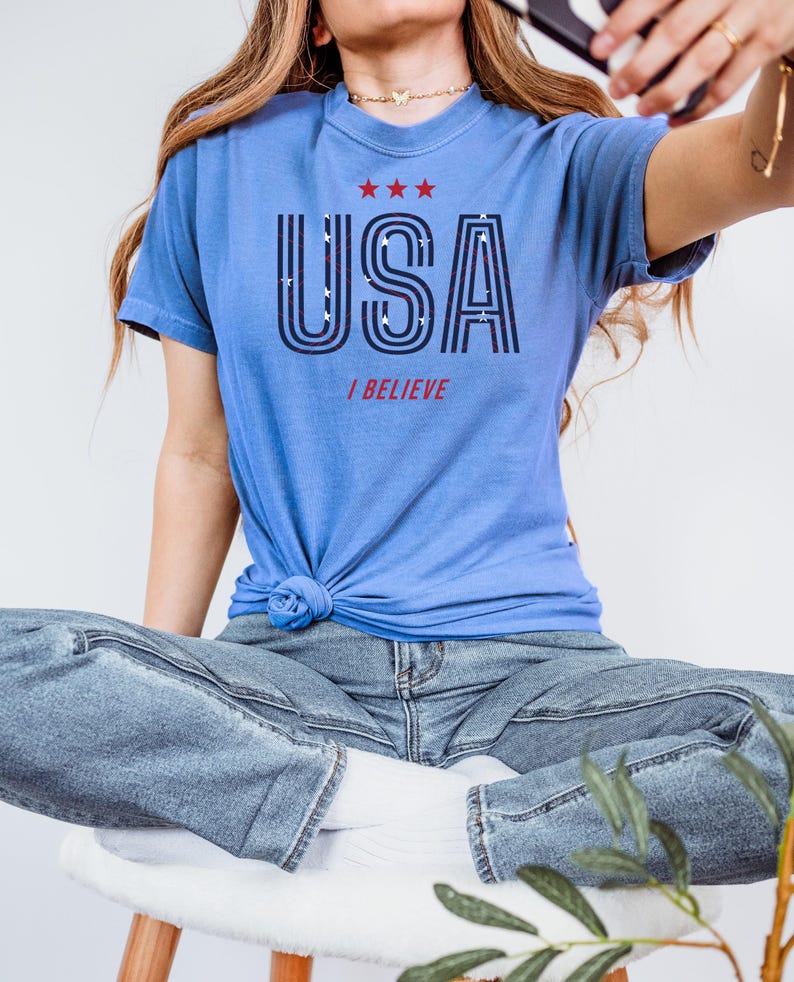 May include: A light blue t-shirt with the word "USA" in a bold, outlined font, and the words "I BELIEVE" below it. The shirt has three red stars above the word "USA". The shirt is knotted at the waist.