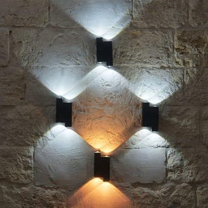 May include: Four rectangular black wall sconces mounted in a diamond pattern on a textured stone wall. Each fixture emits bright beams of light in both upward and downward directions, creating overlapping triangular patterns of illumination.