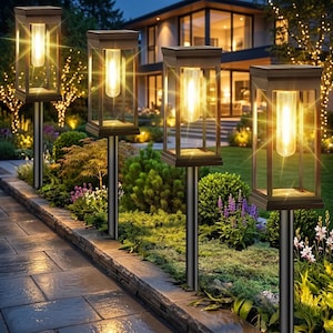 May include: Four rectangular outdoor path lights with dark metal frames and clear glass panels. Each light features a central glowing cylindrical bulb mounted on a vertical post, illuminating a stone walkway and garden area at night.
