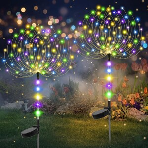 May include: Two solar-powered garden lights with colorful LED lights. Each light has a central stem with a solar panel and a cluster of radiating wires tipped with small, multi-colored lights. The lights are set in a grassy area with a blurred background of plants and bokeh lights.