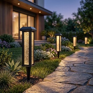May include: A series of black rectangular solar path lights with clear glass panels containing internal bubble textures. Each light features a square top cap and a pointed stake base, positioned along a stone walkway in a garden setting.
