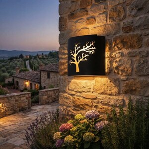 May include: A matte black wall-mounted light fixture with a cutout design of a bare tree. The fixture emits a warm, glowing light from behind the metal panel, casting illumination both above and below the unit against a textured stone wall.