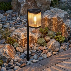 May include: A black solar-powered garden light with a rectangular, vertically ribbed glass shade emitting a warm yellow glow. The fixture is mounted on a thin, straight black metal stake, designed for placement in outdoor landscaping areas.