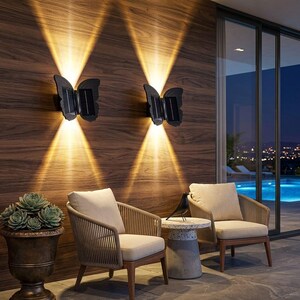 May include: Two black butterfly-shaped wall lights mounted on a wood-paneled wall, each emitting upward and downward beams of warm yellow light. The fixtures feature integrated solar panels on the wings, illuminating the outdoor patio area.