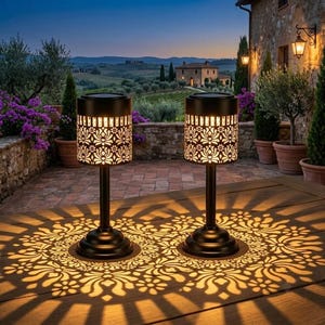 May include: Two black cylindrical solar lamps with intricate floral cutouts stand on a wooden surface. The lamps emit a warm glow, casting detailed, symmetrical, circular light patterns onto the table, creating a decorative shadow effect.