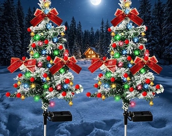 May include: Two artificial Christmas tree garden stakes featuring snow-dusted green branches, red bows, small gold bells, and red berry ornaments. Each tree is topped with a red star and includes integrated solar panels and metal ground spikes.