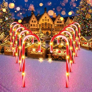 May include: A series of illuminated red and white striped candy cane pathway markers arranged in an archway. Each candy cane features warm yellow LED lights and a hanging star-shaped ornament, set against a festive outdoor holiday market scene.