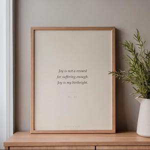 May include: A framed print with the text "Joy is not a reward for suffering enough. Joy is my birthright." The print is in a light wood frame and rests on a wooden surface. A white vase with greenery is to the right.