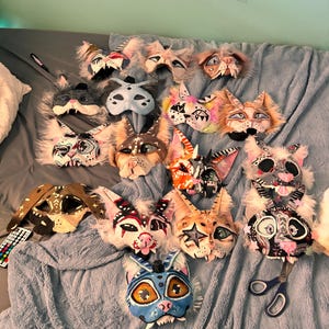 Puede incluir: Una colección de máscaras de gato hechas a mano con diversos diseños y colores, incluyendo blanco, negro, marrón y azul. Algunas máscaras presentan detalles de piel y detalles pintados, con una variedad de expresiones y estilos.