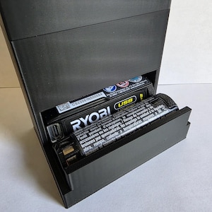 May include: A black rectangular battery storage unit with a sliding drawer. The drawer is partially open, revealing several batteries, including a Ryobi battery and a USB battery. Text is visible on the batteries.