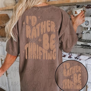 May include: Brown t-shirt with the text "I'd Rather Be Thrifting" in a retro font. The design is in a lighter brown color. A smaller version of the design is in a circle at the bottom right. The shirt is a pocket design.