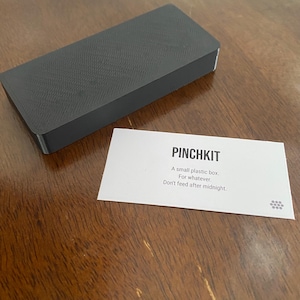 Puede incluir: Una caja rectangular de plástico gris oscuro con una superficie texturizada. Una tarjeta blanca con el texto "PINCHKIT" y las frases "A small plastic box. For whatever. Don't feed after midnight." está al lado de la caja.